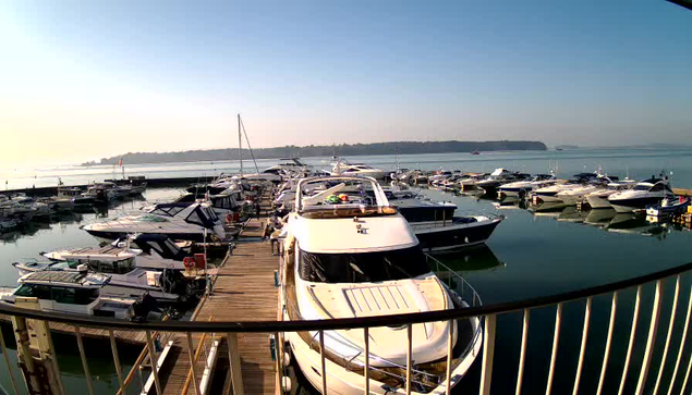 A marina scene featuring multiple boats and yachts docked along a wooden pier. The water is calm and reflects the boats, under a clear blue sky with gentle sunlight. In the background, green hills can be seen along the shoreline.