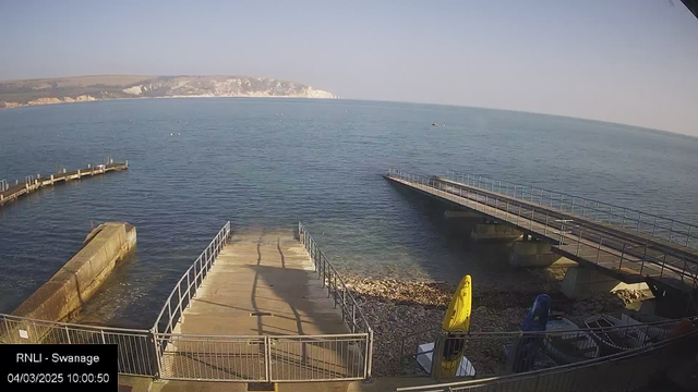 A calm seaside scene captures the water in varying shades of blue, with gentle ripples reflecting the light. On the left, a curved wooden pier extends into the water, while a straight, metal-framed pier is positioned on the right, both leading away from a stone and concrete ramp that slopes down to the water's edge. To the right of the ramp, several kayaks in bright yellow and blue rest on a rocky shore. In the background, white chalk cliffs rise above the water, adding a natural contrast to the seaside landscape. The sky is clear and bright, suggesting a sunny day.