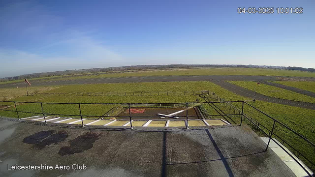 A clear blue sky stretches above a green landscape, featuring a grassy airfield with a runway. In the foreground, there is a railing and a small wooden structure resembling a hangar. A small white airplane is sitting on the ground, near some scattered markings. A windsock is visible in the distance, gently blowing in the breeze. The date and time in the top corner indicate it is 10:01 AM on March 4, 2025.