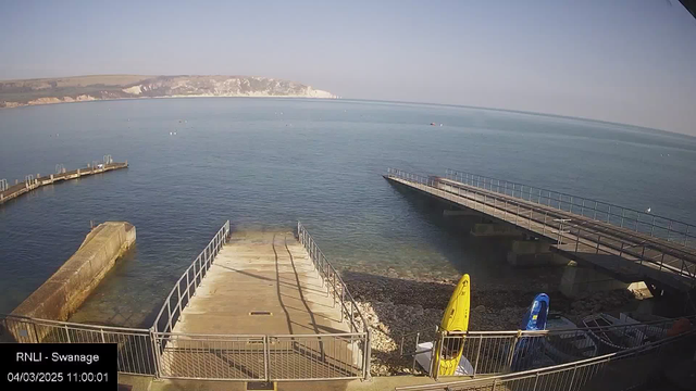 A serene coastal scene featuring calm blue water extending to the horizon. There is a distant white chalk cliff on the left side of the image. Two piers extend into the water from the foreground, with one pier curving to the left. On the right side, there are two kayaks, one yellow and one blue, resting on the shore near the water's edge. The sky is clear and bright, and the image is taken during daytime.