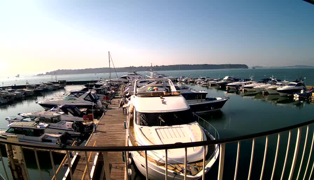 A serene marina scene featuring numerous boats docked in calm waters. The foreground shows a wooden dock with several yachts and smaller boats moored alongside each other. In the background, there are more boats visible on the water, against a clear blue sky. Gentle hills can be seen in the distance, creating a peaceful coastal atmosphere.