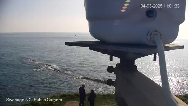 A coastal scene captured from a high vantage point. In the foreground, there is a white object resembling a camera mounted on a metallic stand. Two people stand on the grassy edge near the water's edge, one facing the ocean and the other turned toward them. In the background, the calm sea extends to the horizon, where a small boat is visible. Soft sunlight reflects off the water's surface, and a rocky formation is seen emerging from the sea. The sky is clear, indicating a sunny day.