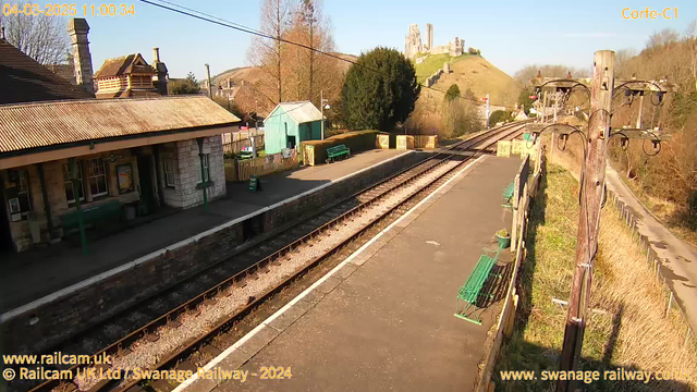 A railway station scene on a sunny day. The foreground features a platform with two parallel sets of railway tracks. There are several green benches and a sign that reads "Way Out." To the left, a stone building with a tiled roof serves as the station house. Behind it, a small blue shed is visible. In the background, a hill rises with a castle ruin on top, surrounded by trees. The sky is clear and blue with a few scattered clouds.