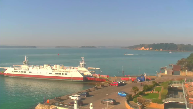 A bright and clear day at a waterfront scene. In the foreground, a large white ferry with red accents is docked at the pier. There are a few parked cars in the area, and a paved surface nearby, with some greenery visible. The water is calm, reflecting the blue sky, and several small boats are moving in the distance. The landscape in the background features lush trees and a sandy shore with cliffs.