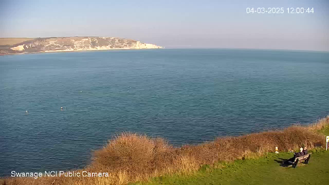 A clear view of a calm sea under a bright blue sky, with gentle waves and a distant coastline characterized by white cliffs. In the foreground, there is a grassy area with a bushy border. Two people are sitting on a bench, facing the water, enjoying the scenic view. The date and time are displayed in the corner.