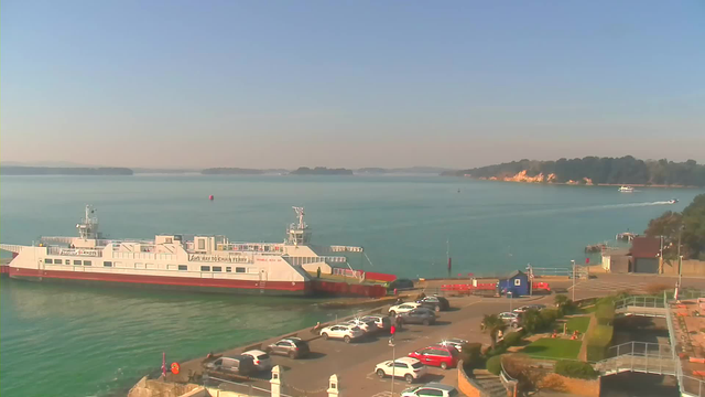 A clear, sunny view of a harbor with a large white and red ferry docked at the pier. In the foreground, there is a parking lot filled with several cars. To the right, there are grassy areas and pathways leading to the water's edge. The background features calm blue water and distant, tree-covered hills. A small boat is visible in the water, and the sky is bright with few clouds.