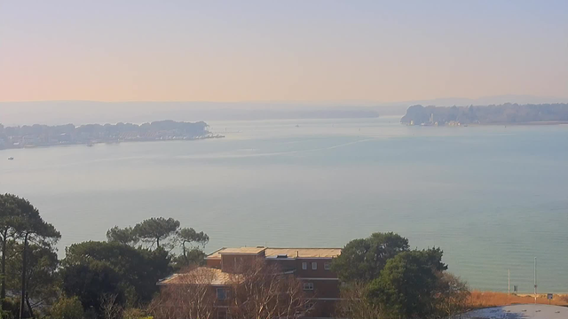 A serene water view is captured, showcasing a calm body of water extending into the distance with a soft hazy atmosphere. The far shore features tree-lined land and a few buildings, while several small boats can be seen dotting the water. In the foreground, there is a mix of trees and a low-rise building with a flat roof, surrounded by other greenery. The overall scene conveys a peaceful coastal landscape under a clear sky with subtle hues.