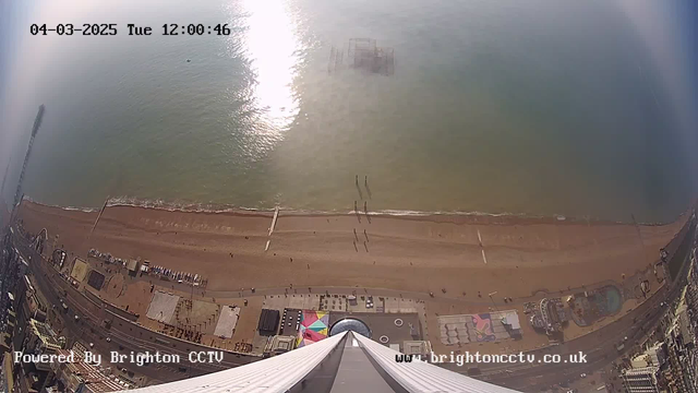 A bird's-eye view of a sandy beach with a shallow, blue-green sea. The coastline is lined with people walking, and shadows are cast on the wet sand. In the distance, a structure submerged in the water, likely a pier, is visible. The left side of the image shows a tall building or structure, with a road and various colorful attractions below, including a circular area and a swimming pool. The sky above is bright with sunlight reflecting off the water. The timestamp indicates it is Tuesday, March 4, 2025, at noon.