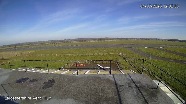 A view from a high vantage point overlooking an airfield. In the foreground, there is a flat surface with some dark patches and horizontal white lines. Beyond that, a runway stretches across the center of the image, bordered by green grass. In the distance, there are hills and a blue sky with some wispy clouds. A red windsock is visible on the left, and wooden fencing outlines the airfield area.