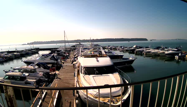 A view of a marina filled with multiple boats of various sizes docked in a calm body of water. The scene is bathed in bright sunlight, with a clear blue sky above. In the foreground, a larger white boat is prominently displayed, surrounded by smaller vessels. The docks extend out into the water, showcasing a busy recreational area. In the background, a distant landmass can be seen, likely an island or peninsula. The overall atmosphere is peaceful and inviting, typical of a seaside setting.