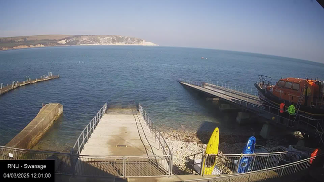 A clear view of a calm sea under a blue sky. In the foreground, there are wooden piers extending into the water, with a rocky shoreline nearby. Two brightly colored kayaks, one yellow and one blue, are stationed on the shore. On the right, a lifeboat is docked at the pier, and a few people in protective gear are visible near the boat. The scene conveys a tranquil coastal environment.