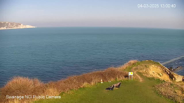 A scenic view of the sea from a coastal area. The horizon features calm blue waters under a clear sky, with a slight gradient of color from light blue near the skyline to deeper blue closer to the water. In the foreground, there is a grassy area with a bench facing the sea. To the right, a pathway leads down towards the water, and there are some shrubs and bushes along the edge. A caution sign is visible nearby. The overall impression is serene and natural, capturing a peaceful coastal landscape.