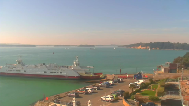 A coastal scene featuring a ferry docked at a terminal. The water is a calm turquoise, with several other boats visible in the distance, including sailboats. The shoreline is lined with greenery, and there is a parking lot filled with cars in the foreground. The sky is clear with a few clouds, suggesting a sunny day.