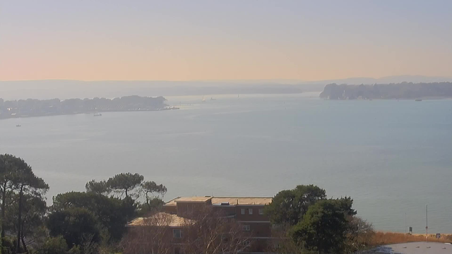 A serene view of a body of water, likely a bay, with gentle ripples reflecting soft pastel colors of a hazy sky. In the foreground, there are trees and part of a building with a flat roof, while in the distance, boats are visible dotting the water. The opposite shore is lined with trees and structures, partly obscured by the misty atmosphere, indicating a tranquil coastal scene.