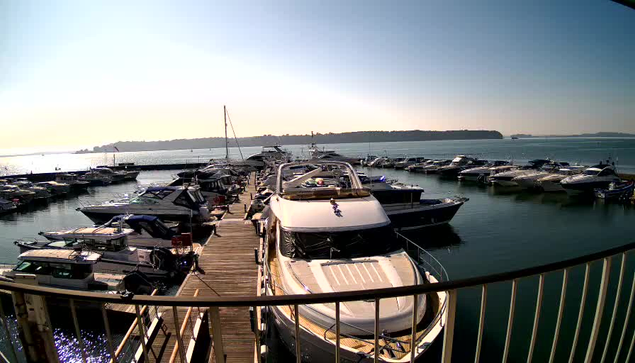 A scenic view of a marina filled with numerous boats docked in calm waters. The foreground features a large white yacht with a sun deck, while several smaller boats are seen alongside. A wooden walkway runs along the dock, providing access to the vessels. In the background, a clear blue sky meets the horizon where the water reflects light, and distant land is visible. The overall atmosphere suggests a sunny day at the waterfront.