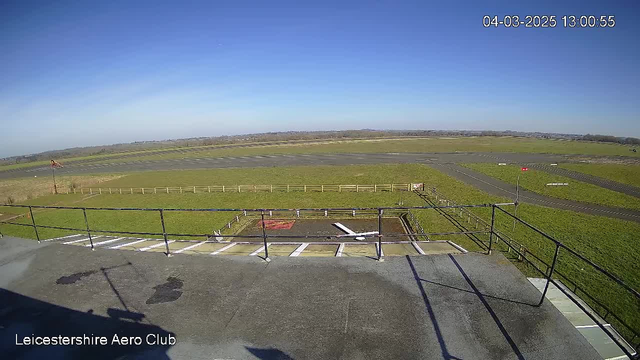 A wide view from a webcam shows a clear blue sky above a green field and a runway. In the foreground, a partial rooftop ledge and railing can be seen. There are markers on the runway and a row of wooden fencing separating the runway from the grass area. In the distance, hills are visible under the vast sky. The scene appears to be at the Leicestershire Aero Club, with no aircraft present at the moment.