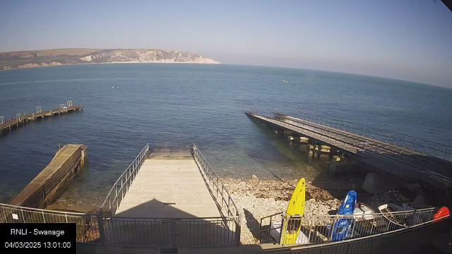 A seaside view featuring a calm blue ocean under a clear sky. In the foreground, there are two wooden jetties extending into the water. On the left, a stone structure leads to a slightly elevated wooden platform. A collection of colorful kayaks in yellow, red, and blue are parked on a rocky shore, with pebbles and some seaweed visible. The distant landscape includes rocky cliffs and hills along the shoreline.