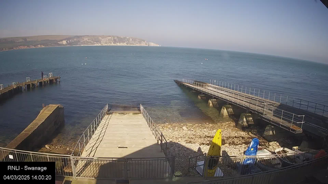 A coastal scene showing a calm sea with a clear blue sky. In the foreground, there is a wooden jetty extending into the water, with several small boats moored nearby. A man stands on the jetty, looking out over the water. To the right, another jetty is visible, also leading into the sea. On the shore, there are kayaks in yellow and blue, and rocky ground scattered with pebbles. In the distance, a cliff formation is visible along the shoreline, providing a natural backdrop to the tranquil setting.