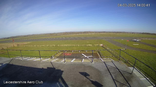 A clear blue sky is visible above a grassy airfield with a taxiway that runs horizontally across the middle of the image. In the distance, a small airplane is parked on the runway to the right. To the left, there is a wooden fence bordering the airfield. The foreground features a railing from a raised platform, along with ground markings that indicate the layout. The image is captured around 2 PM on March 4, 2025.