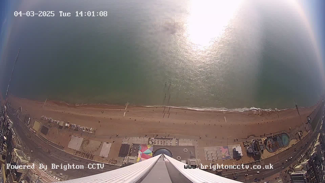 A high-angle view of a beach and coastline, captured from a webcam. The image shows a sandy beach with people walking and a few scattered colorful beach umbrellas. There is a body of water in the foreground reflecting sunlight. A pier is visible in the water, and several buildings and structures are positioned along the beach. The date and time are displayed in the top left corner, and the image is tagged as powered by Brighton CCTV.