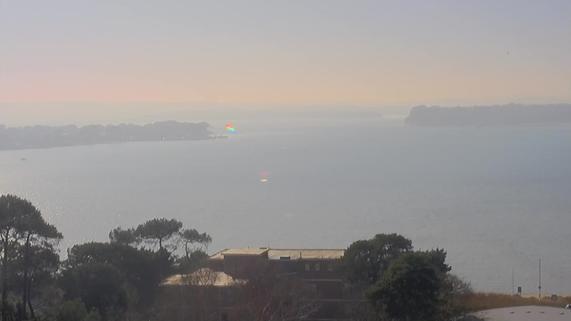 A serene coastal view featuring a calm body of water stretching into the distance. The horizon displays soft pastel colors, suggesting a morning or evening glow. In the foreground, there are trees with dark green foliage. A few buildings with flat roofs are visible, partially obscured by branches. A small boat is seen on the water, and a subtle rainbow effect glimmers along the shoreline. The scene conveys a tranquil and picturesque atmosphere.