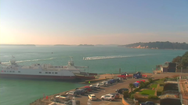 A scenic view of a harbor during the daytime. In the foreground, there is a large ferry boat docked at the pier, labeled with a sign that reads "GIVE WAY TO CHAIN FERRY." Several cars are parked along a paved area near the water. In the background, the sea is a bright turquoise, with a few small boats creating wake as they navigate the water. Beyond the water, hills can be seen in the distance, along with tree-covered areas and some rocky cliffs. The sky is clear with a light gradient from blue to soft peach near the horizon.