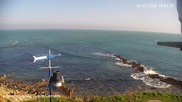 A coastal scene captured by a webcam shows calm blue waters stretching toward the horizon, with gentle waves lapping against rocky outcrops on the shore. To the left, there is a weather vane with a stylized fish design, mounted on a blue pole above a black cylindrical object. The foreground features a mix of rocky terrain with sparse vegetation, while the sky maintains a clear and sunny appearance. A timestamp in the corner indicates the date and time of the image.