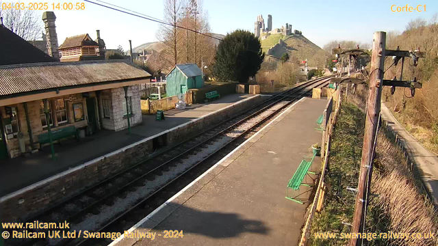 A view of Corfe Castle station on a clear day. The foreground features a stone platform with two sets of railway tracks. There are wooden benches along the platform and a small green building visible in the background. To the right, a wooden post with electrical wires runs parallel to the tracks. In the distance, Corfe Castle is perched on a hill, surrounded by trees and greenery. The time and date are displayed in the top left corner.