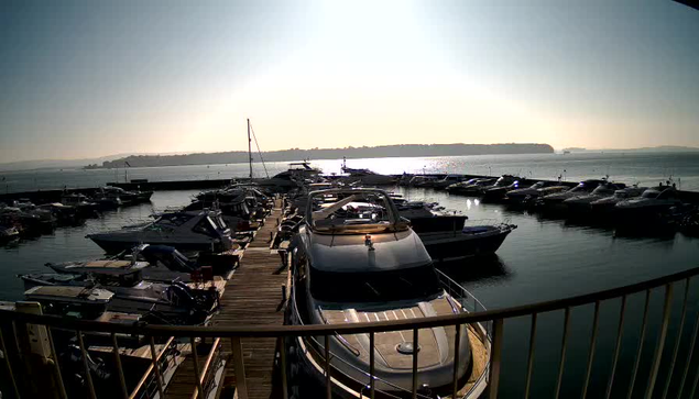 A marina scene under bright sunlight, with numerous boats docked in calm water. The foreground features a large white boat with a shiny, curved surface. Both sides are lined with smaller vessels of various sizes and colors, while the background shows a distant landmass and a clear sky. Reflection of the sun glimmers on the water, creating a serene atmosphere.