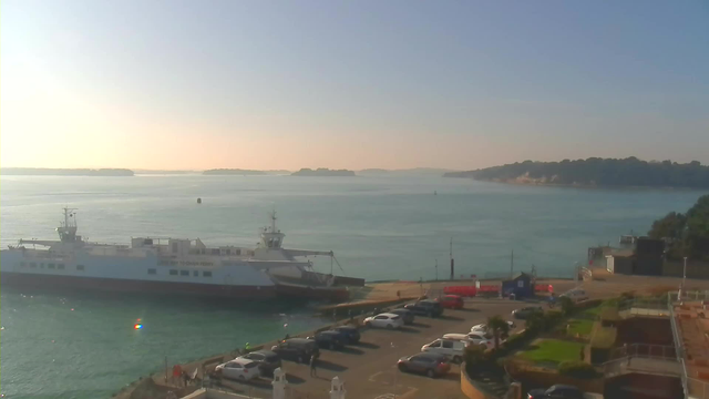 A calm harbor scene features a large white ferry docked at a pier. In the foreground, a parking lot is filled with various parked vehicles, including cars and a few people walking. The water is a gentle turquoise, with distant islands visible against a pale sky. The sun is low on the horizon, casting a warm glow on the scene. To the right, a hillside is covered with greenery, and a small building can be seen. The overall atmosphere is serene and inviting.