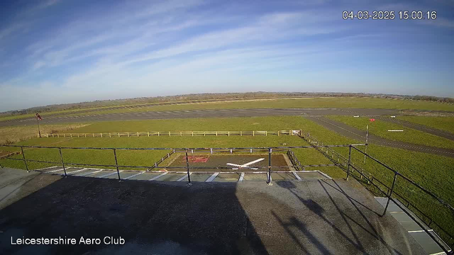 A view from a webcam positioned on a building overlooking a grassy airfield. In the foreground, there is a railing along the edge of the building. Below, a runway marked by a white cross and various lane markings can be seen. The airfield is surrounded by a wooden fence, and in the background, there are low green hills and blue sky with some clouds. The image has timestamp details indicating the date and time.