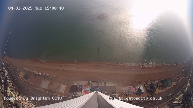 An aerial view of a beach scene taken from a high vantage point. The image shows a sandy shoreline meeting calm blue water, with small waves lapping at the shore. Along the beach, there are distant figures of people and several structures, including a pier and colorful beach huts. The scene is bathed in sunlight, creating glimmers on the water's surface. The date and time are displayed in the top left corner.