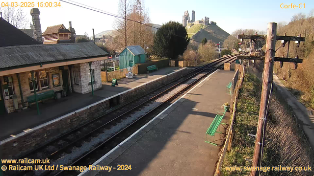 A view of a train station during the day with clear blue skies. On the left, there is a small stone building with a sloped roof, featuring several windows and a green bench in front. There is a green shed behind the station building. The platform is occupied by a few green benches and a small sign that reads "WAY OUT." Train tracks run along the bottom of the image, leading towards the distance. In the background, a hill rises with the remains of a castle on top. The area is bordered by trees and hedges, creating a scenic landscape.