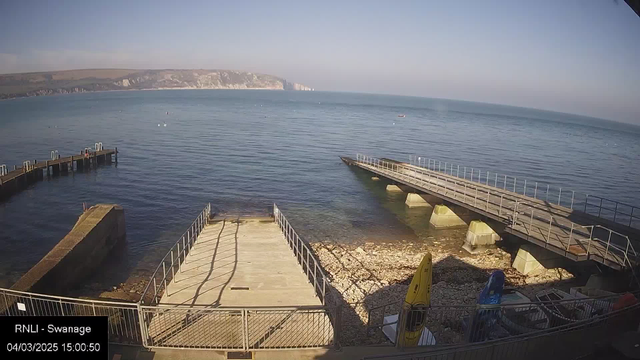 A coastal scene showing a clear blue sea under a bright sky. In the foreground, there is a concrete ramp leading down to the water, with a rough, rocky shoreline to the left. On the right, two piers extend into the water, with one being straight and the other curved. A few boats are moored nearby. The background features distant cliffs, hinting at a scenic landscape. The time and date are displayed in the bottom left corner.