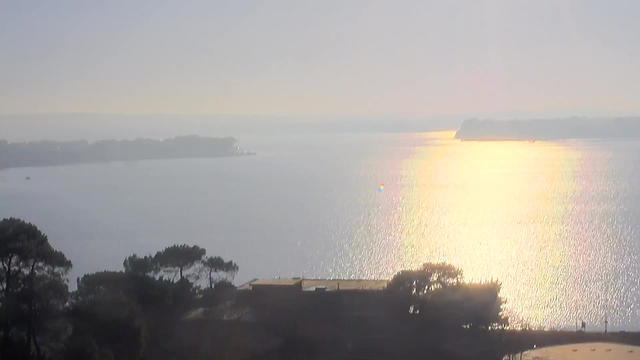 A tranquil view of a calm body of water reflecting sunlight, creating a shimmering effect. In the foreground, there are dark silhouettes of trees and a low building. In the background, a distant coastline is visible, merging with a hazy sky. The overall scene conveys a peaceful and serene atmosphere.