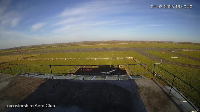 A wide view of an open airfield on a sunny day. The foreground shows a flat rooftop with a railing, and a large white "X" marking on a tarmac area, which is likely a helipad or landing zone. In the background, green grass stretches across the airfield, with a few white fences outlining the perimeter. There are two small red flags on poles, indicating the wind direction. The sky is mostly clear with some blue and wispy clouds.