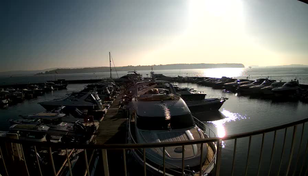 A marina filled with various boats and yachts docked in calm water. The sun rises in the sky, illuminating the scene and casting a reflection on the water's surface. In the background, hills are visible, creating a scenic coastal view. The atmosphere appears tranquil and inviting.