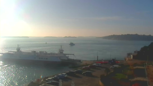 A tranquil waterscape features a large white ferry docked in the foreground with a smaller boat navigating the water nearby. The sun casts a warm glow over the scene, illuminating the gentle waves. In the background, a series of green hills or islands rise from the water, while a parking area with several cars is visible along the shoreline. The sky transitions from blue to gold, indicating early morning or late afternoon.