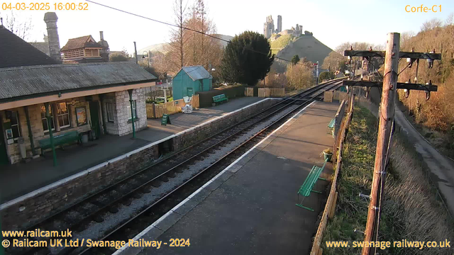 A view of Corfe Castle railway station on a clear day. The station features a stone platform with green benches on either side. To the left, there is a building with a sloped roof and a chimney. A small, blue structure is visible further along the platform. The railway tracks run straight through the station, and in the background, Corfe Castle sits atop a hill, partially obscured by trees. The scene captures a tranquil atmosphere with bright sunlight illuminating the landscape.