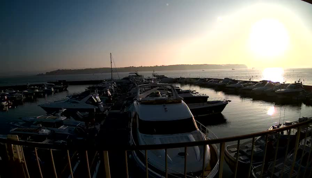 A serene marina scene at sunrise, with numerous boats docked in calm waters. The sun is partially visible on the horizon, casting a warm glow across the water, creating reflections. In the background, gentle hills are outlined against the brightening sky, which transitions from deep blue to soft golden hues. A railing is visible in the foreground, framing the scene.