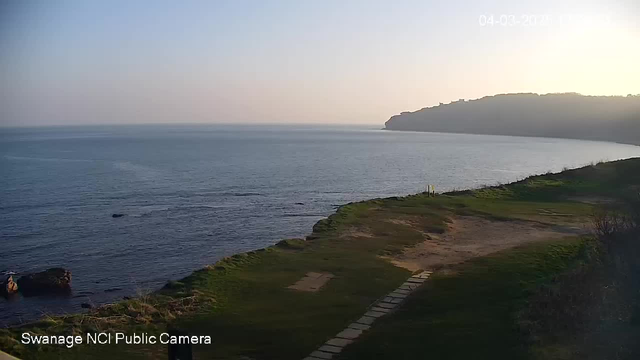 A scenic view of the sea at sunrise or sunset, with calm blue water extending to the horizon. The shoreline is visible in the foreground, featuring grassy areas and a path made of stone or concrete. There are a few dark rocks along the shore. In the distance, a gentle rise of land forms a cliff. The sky has a light color gradient, with soft hues transitioning from light blue to warmer tones near the horizon. The image is marked with a timestamp indicating the date and time, along with a label for the webcam.