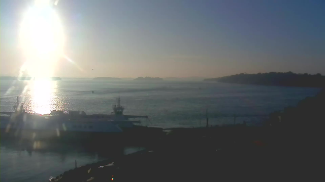 A bright sun glows in the sky, reflecting off the calm waters of a bay. A silhouette of a large ship is visible in the foreground, docked at a pier. In the background, distant islands and a shoreline are faintly seen under a clear blue sky, creating a serene coastal scene.