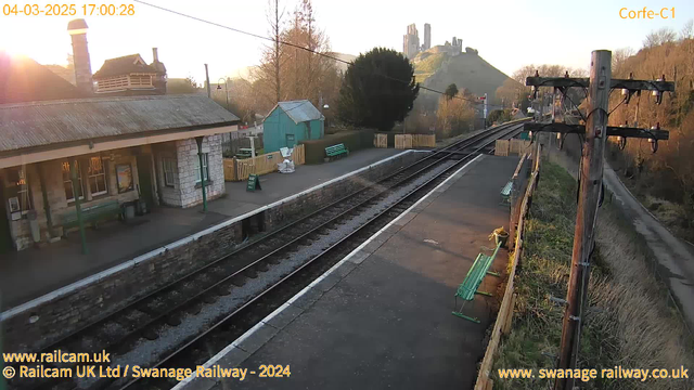 A railway station scene during sunset. The foreground shows a platform with two green benches and a stone wall. In the background, there is a small building with a sloped roof and large windows. To the right, there are train tracks leading toward the horizon. On a hill in the distance, ruins of a castle can be seen, silhouetted against the sunset sky. Trees and a fence border the scene, with the sun casting a warm glow over the area.