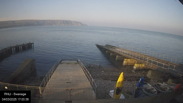 A calm coastal scene is depicted, featuring a still body of water reflecting the light of the sky. In the foreground, there are two piers extending into the water, with one pier having a ramp leading to a lower area of rocky shore. Yellow and blue kayaks are positioned on the side of the pier, and small boats can be seen anchored in the water. The coastline in the background shows a rocky linear formation under a clear sky.