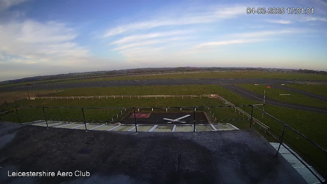 A clear view of an airfield taken from a high vantage point. In the foreground, there is a section of a building with a roof and railing. Below the railing, a runway is visible, accompanied by a large open field filled with grass. A white aircraft landing strip marking is positioned in the field, while a red indicator can be seen in the distance. The sky is mostly clear with some scattered clouds, and the scene is well-lit, suggesting daylight.