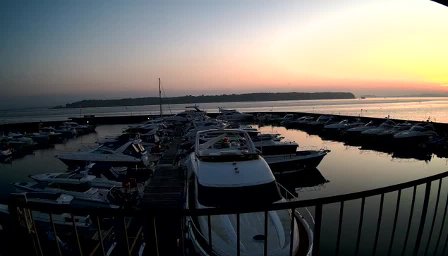 A marina is depicted at dawn, with numerous boats docked in the water, reflecting the soft orange and pink hues of the rising sun. The scene includes a distant shoreline with trees silhouette and a calm water surface. The boats vary in size and design, some are white, while others are darker. A railing is visible in the foreground, framing the scene.