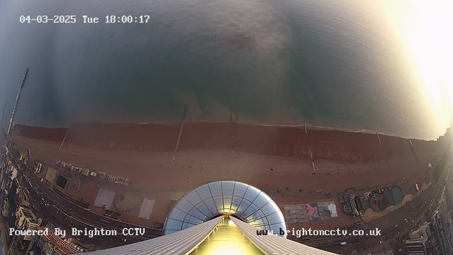 Aerial view from a high vantage point overlooking a beach. The camera captures a wide stretch of sandy shore meeting the calm sea. The beach is lined with a pier extending into the water on the left side. In the foreground, the observation platform features a circular glass structure with a glowing edge. The image shows a blend of sand, water, and part of the coastal road with some buildings and attractions visible along the beach. The upper corner displays a timestamp indicating the date and time of the capture.