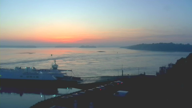 A calm sea at sunrise, with hues of orange and pink reflecting on the water. A ferry is docked at the foreground, partially illuminated. In the distance, there are silhouettes of land and trees along the shoreline. The sky transitions from light blue to darker shades, indicating the early morning hour. Some vehicles are parked near the waterfront.