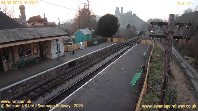 A view of a railway station platform with multiple green benches along the edge. The building on the left features a stone facade and a sloped roof, with a sign indicating "Corfe Castle." In the background, a hill is visible with the ruins of Corfe Castle at the top. The platform has two railway tracks, leading out of the frame, with a signal post on the right and some greenery beyond the tracks. The scene is set during the evening, as indicated by the soft lighting.