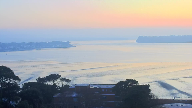 A serene waterfront scene at dawn, featuring calm water reflecting soft pastel colors of pink, orange, and blue in the sky. In the foreground, there are silhouettes of trees and a building with dark hues. The distant shoreline is lined with houses, and a small boat can be seen on the water. The overall atmosphere is tranquil and peaceful, suggesting early morning light.